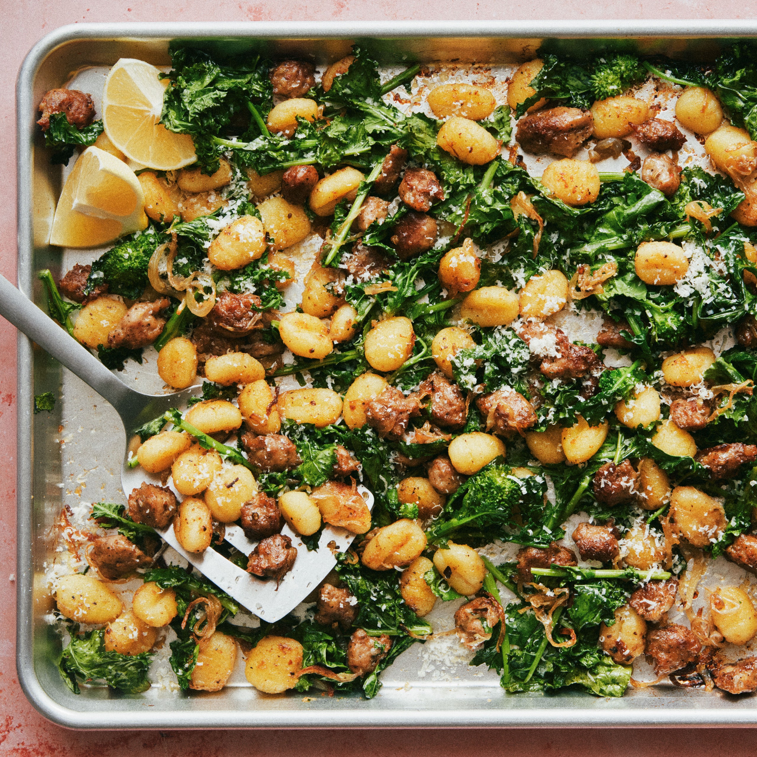 Gnocchi on a sheet tray with pieces of Italian sausage broccoli rabe red onions parmigiana and lemon wedges.